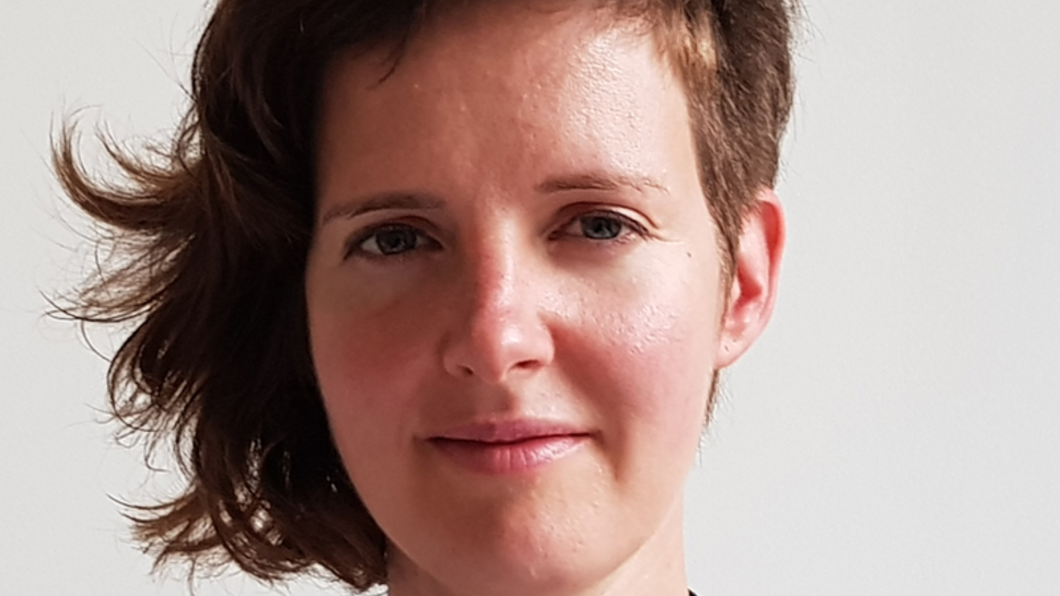 Foto van Rinske Wessels, een witte vrouw met bruin haar. Ze is vanaf haar schouders naar boven in beeld, heeft een zwart jasje aan en is gefotografeerd op een lichtgrijze, neutrale acthergrond.